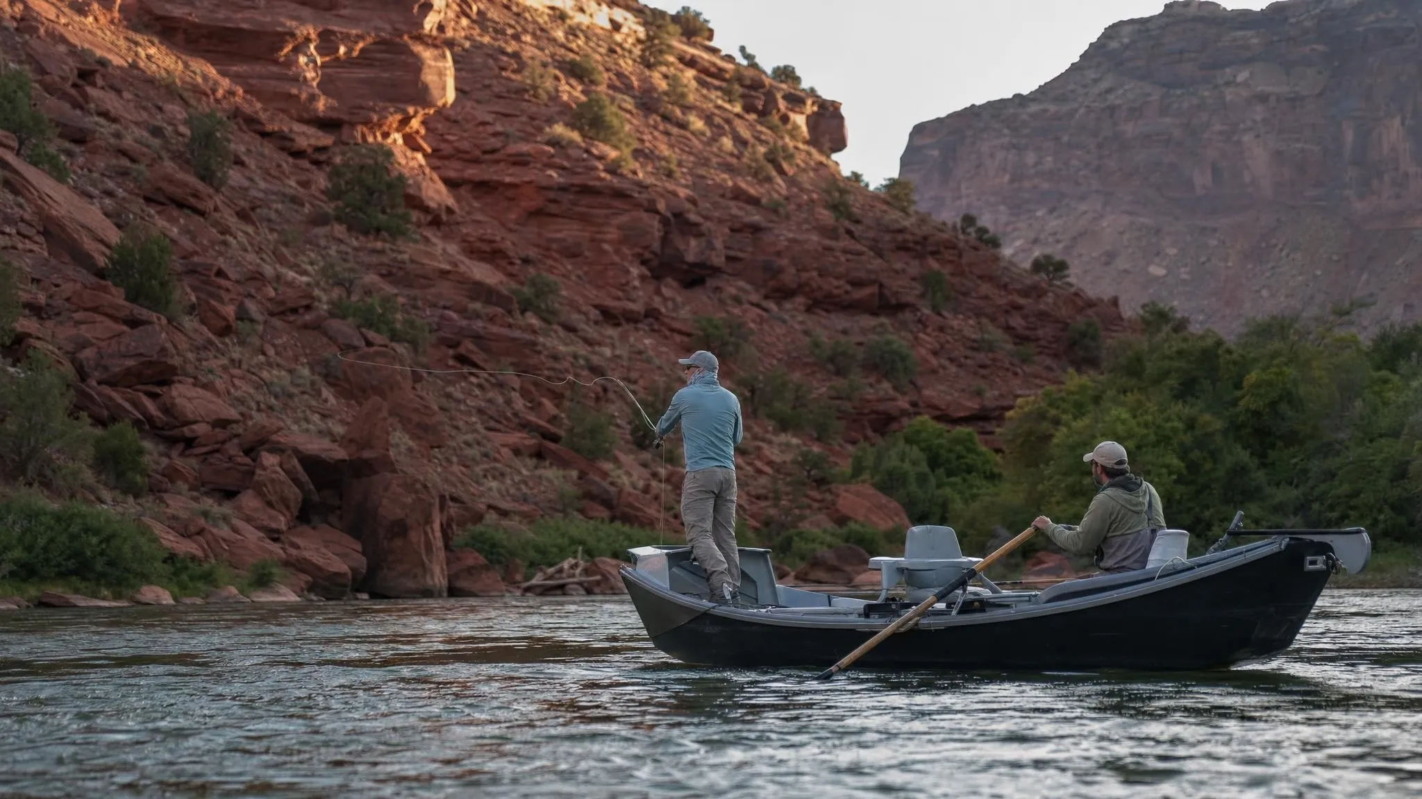 A wooden drift boat on a green river in a red-rock canyon, a guide rowing while an angler casts from the bow